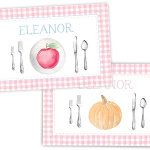 Op de afbeelding: Twee roze en witte gingham placemats met de naam "Eleanor" erop gedrukt. De bovenste placemat heeft een rode appel op een wit bord met een vork, mes en lepel. De onderste placemat heeft een gele pompoen met een vork, mes en lepel.