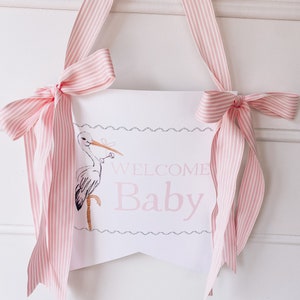 Może przedstawiać: Biały baner z r&oacute;żowym napisem "Welcome Baby" i r&oacute;żową wstążką w białe paski wiszącą u g&oacute;ry. Po lewej stronie banera znajduje się ilustracja bociana.