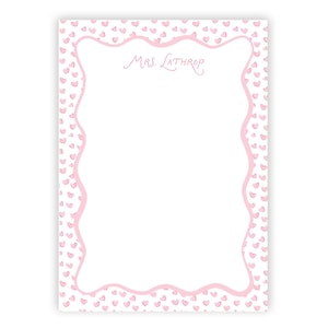 Può includere: Un blocco note rettangolare con sfondo bianco e motivo a cuori rosa. Il bordo è una linea rosa ondulata. Il blocco note riporta la scritta "MRS. LATHROP" in corsivo rosa.