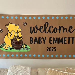 May include: A brown banner with a Winnie the Pooh illustration, bees, and the text "welcome BABY EMMETT 2025". The banner has a blue dotted border and is hanging on a wall.