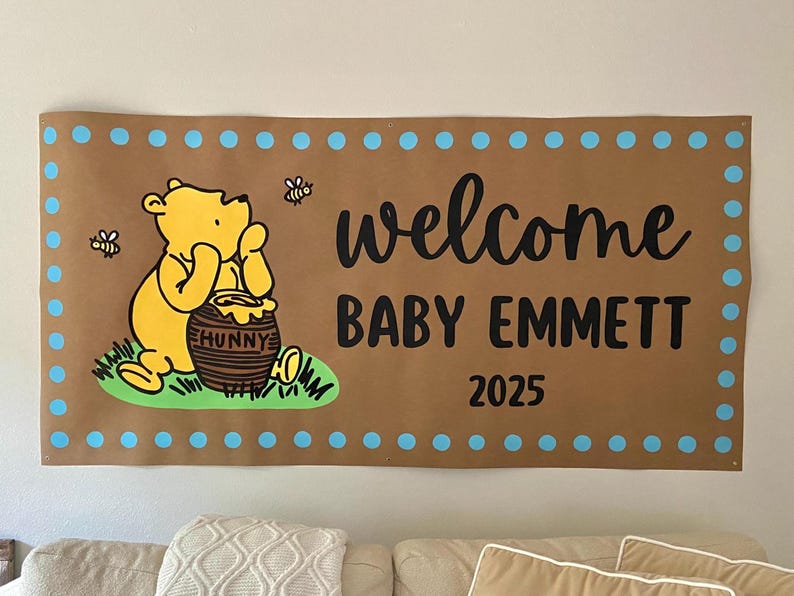 May include: A brown banner with a Winnie the Pooh illustration, bees, and the text "welcome BABY EMMETT 2025". The banner has a blue dotted border and is hanging on a wall.