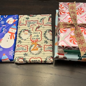 May include: Three wrapped Christmas gifts with festive paper. The first gift is blue with a snowman design, the second is beige with a reindeer design and the text "Have a Jolly Holiday", and the third is pink with a candy cane design.