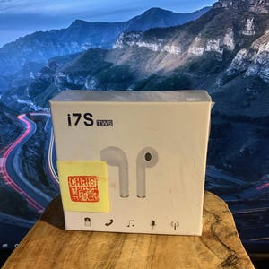 May include: White box of i7S TWS wireless earbuds with a pair of white earbuds. The box has the text "i7S TWS" and icons. A yellow sticker with red text is on the box. The background shows a mountain scene.