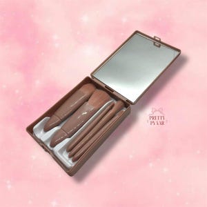 May include: A brown makeup brush set with a mirror in a compact case. The case has a white interior with slots for the brushes. The brushes are made of synthetic bristles and have brown handles.