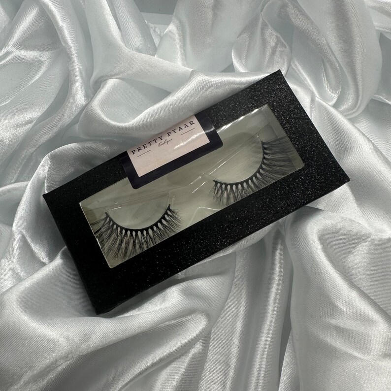 K&ouml;nnte beinhalten: Eine schwarze Box mit einem durchsichtigen Fenster, in dem zwei Paar falsche Wimpern zu sehen sind. Auf der Box steht "PRETTY PYAAR".