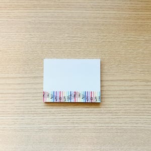 May include: A white rectangular notepad with a colorful striped border and musical notes. The border features vertical stripes in various colors, including blue, pink, and yellow. Black musical notes are printed over the stripes.