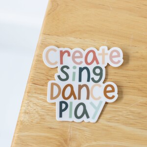 May include: A white sticker with the words "create", "sing", "dance", and "play" in colorful, stylized lettering. The sticker is on a light brown wooden surface.