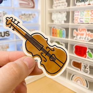 May include: A brown and black violin sticker with a watercolor effect. The sticker is being held by a hand.