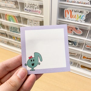 May include: A purple sticky note with a light blue musical note character with a red bow. The character has a smiling face and is holding its arms out. The note is on a wooden surface.