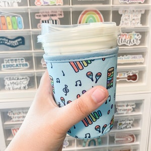 Peut inclure: Une manchon de tasse réutilisable bleu clair sur le thème de la musique. Le manchon présente des notes de musique, un clavier de piano arc-en-ciel et des maracas. Le manchon est sur une tasse blanche avec un couvercle transparent.