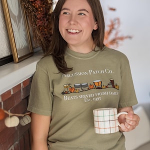 Könnte beinhalten: Ein olivgrünes T-Shirt mit einer Grafik aus verschiedenen Schlaginstrumenten und Kürbissen. Der Text lautet Percussion Patch Co., Beats Served Fresh Daily und Est. 1995. Eine weiße Tasse mit farbigem Karomuster wird im Vordergrund gehalten.