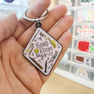 Könnte beinhalten: Ein pinkfarbener und gelber Emaille-Pin in Form einer Raute mit dem Text "BAND ROOM Keys" und einem Smiley. Der Pin zeigt Illustrationen von Musikinstrumenten, darunter eine Klarinette, eine Trompete, ein Posaune, eine Flöte, eine Trommel und ein Saxophon.