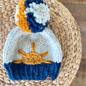 May include: A knitted hat with a pom pom on top. The hat is white with a blue and yellow stripe at the bottom. The hat has a yellow sun design on the front. The hat has a brown leather tag with the text "Happy Trails" on it.