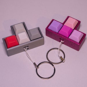 May include: Two Tetris-shaped keychains, one gray and one pink, with different colored keys. Each keychain has a silver split ring.