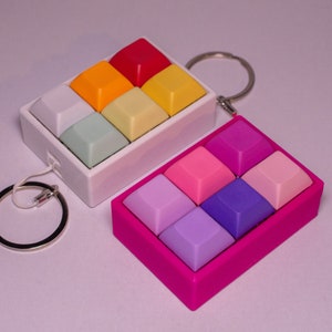 Könnte beinhalten: Zwei Schlüsselanhänger mit bunten Tastenkappen. Der erste Schlüsselanhänger ist weiß mit sechs Tastenkappen in Regenbogenfarben. Der zweite Schlüsselanhänger ist pink mit sechs Tastenkappen in Violett- und Rosatönen.
