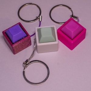 Könnte beinhalten: Drei Schlüsselanhänger mit unterschiedlich farbigen Tastenkappen. Die Tastenkappen sind lila, grün und pink. Jeder Schlüsselanhänger hat einen silbernen Ring und eine silberne Kette.