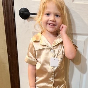 May include: A young child wearing a gold satin pajama set with short sleeves and shorts. The child is standing in front of a white door with a black door handle.
