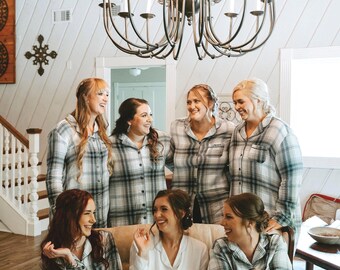 May include: A group of women wearing matching plaid pajama sets, some seated on a beige sofa. One woman wears a white pajama set with embroidered initials. A large chandelier hangs above them.