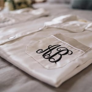 May include: A white button-down pajama shirt with a folded collar and a chest pocket. The pocket features a black embroidered monogram with the letters AB in a cursive script. The shirt is made of a smooth, light-reflecting fabric with long sleeves.