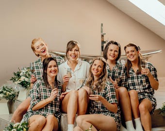 May include: A group of people wearing matching green and white plaid pajama sets, sitting on steps. One person wears a white button-down shirt. Each person holds a champagne flute. White flowers in square vases are on the steps.