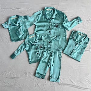 May include: A set of four teal blue satin pajamas with black trim and personalized embroidery. The set includes a long-sleeved shirt, pants, and two short-sleeved shirts.