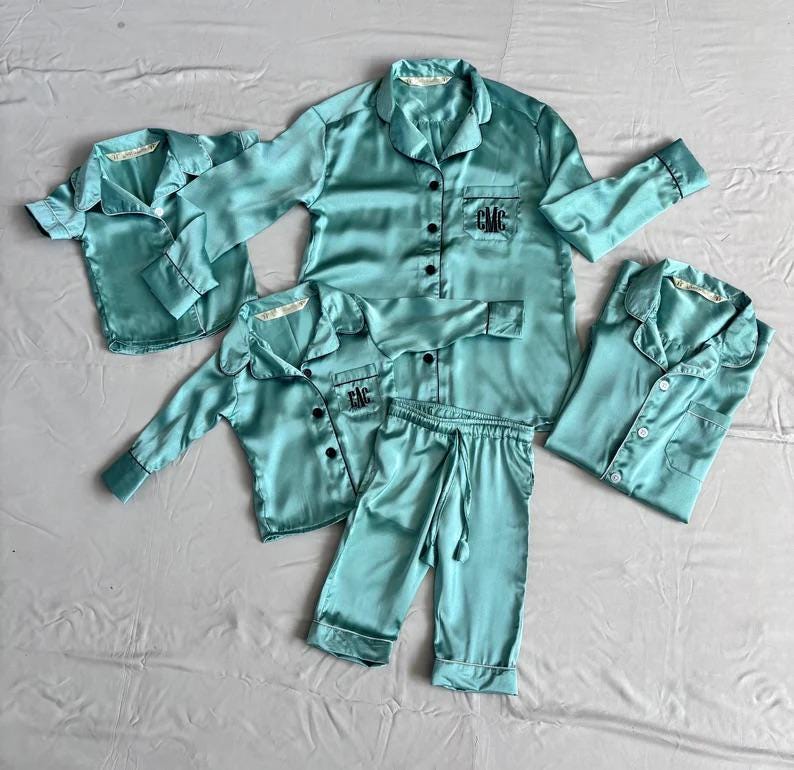 May include: A set of four teal blue satin pajamas with black trim and personalized embroidery. The set includes a long-sleeved shirt, pants, and two short-sleeved shirts.