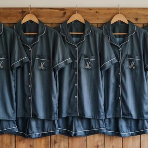 Pode incluir: Cinco conjuntos de pijama azul escuro a condizer pendurados em cabides de madeira. Cada conjunto inclui uma camisa de manga curta com bot&otilde;es e cal&ccedil;&otilde;es, com debrum branco. O bolso da camisa apresenta uma letra "M" bordada.