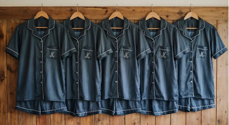 Pode incluir: Cinco conjuntos de pijama azul escuro a condizer pendurados em cabides de madeira. Cada conjunto inclui uma camisa de manga curta com bot&otilde;es e cal&ccedil;&otilde;es, com debrum branco. O bolso da camisa apresenta uma letra "M" bordada.