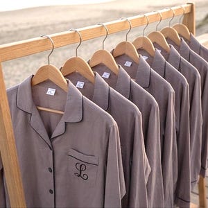 Puede incluir: Una fila de camisas abotonadas de color topo con ribetes negros y una inicial bordada en el bolsillo, colgadas de perchas de madera. Las camisas se exhiben en un estante de madera, con una escena de playa al fondo.