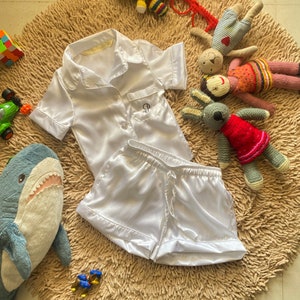 Puede incluir: Conjunto de pijama de satén blanco con mangas cortas y pantalones cortos. La parte superior tiene cierre de botones y un bolsillo con un monograma en el pecho izquierdo.