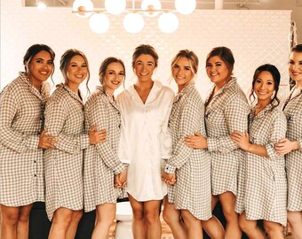 May include: A group of women wearing matching gray and white checkered button-down shirts and a white satin shirt. The women are standing close together, smiling, and holding hands. The setting appears to be indoors, with a decorative light fixture in the background.