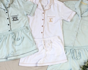 May include: Three sets of pajamas in the image. Two sets are light blue, and one is white. Each set includes a short-sleeved button-down shirt and matching shorts. The shirts have contrasting trim and embroidered initials on the pocket. Wooden hangers are in the background.