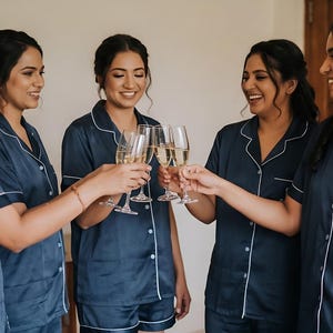 Pode incluir: Cinco mulheres em conjuntos de pijama azul marinho combinando com acabamento branco, brindando com ta&ccedil;as de champanhe. Os pijamas t&ecirc;m uma camisa com bot&otilde;es e shorts. Uma camisa tem um monograma. As mulheres est&atilde;o sorrindo e parecem estar celebrando.