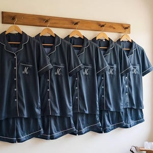 Pode incluir: Seis conjuntos de pijama azul marinho pendurados em um cabide de madeira. Cada conjunto inclui uma camisa de manga curta com bot&otilde;es e cal&ccedil;&otilde;es, com debrum branco. As camisas s&atilde;o bordadas com uma inicial branca.