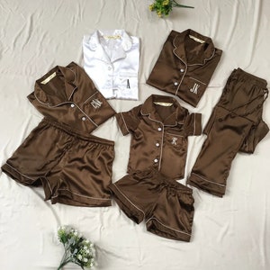 May include: A set of brown satin pajamas with white piping. The set includes a short-sleeved top, long pants, and shorts. The tops have a button-up front and are monogrammed with the letters "A", "JK", and "FK".