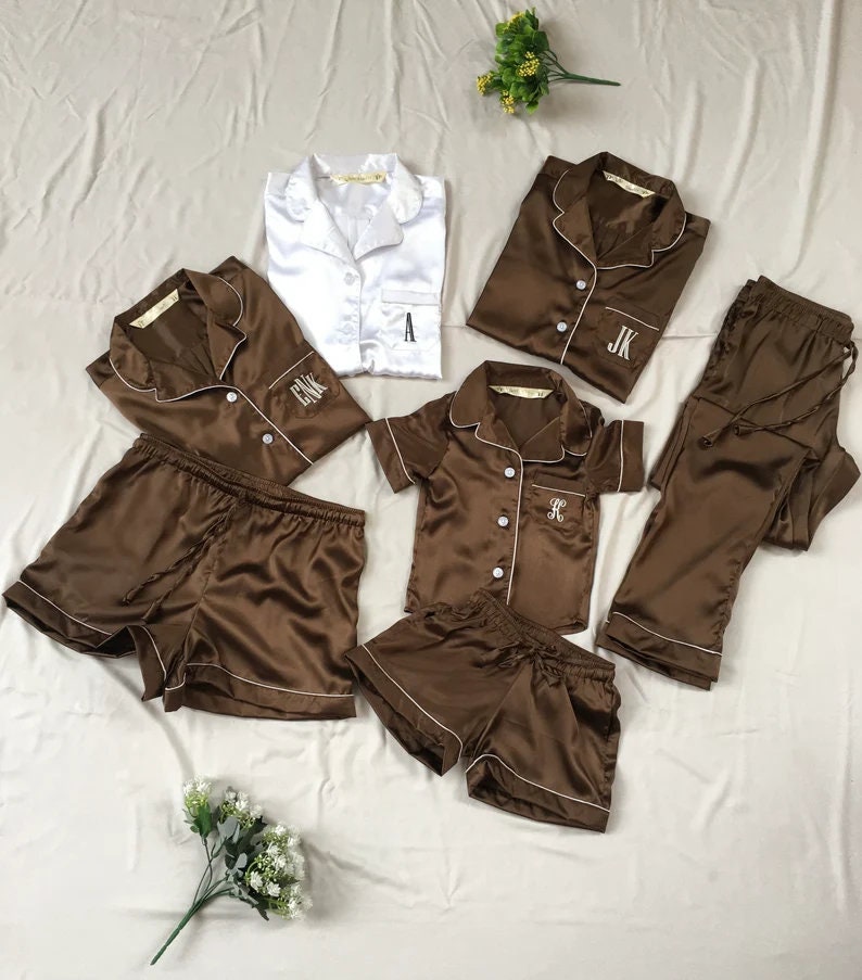 May include: A set of brown satin pajamas with white piping. The set includes a short-sleeved top, long pants, and shorts. The tops have a button-up front and are monogrammed with the letters "A", "JK", and "FK".