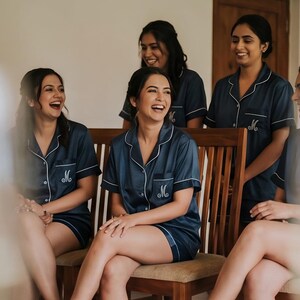 Pode incluir: Um grupo de mulheres vestindo conjuntos de pijama azul marinho combinando com acabamento branco e a letra "M" bordada no bolso. Elas est&atilde;o rindo e sentadas em uma cadeira de madeira. A cena &eacute; bem iluminada.
