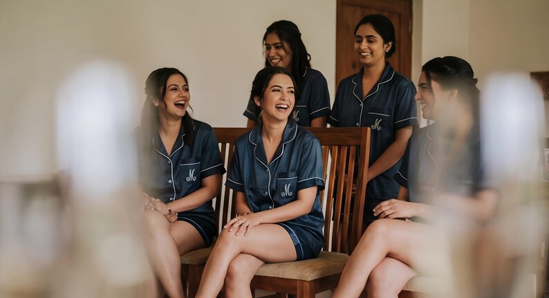 Pode incluir: Um grupo de mulheres vestindo conjuntos de pijama azul marinho combinando com acabamento branco e a letra "M" bordada no bolso. Elas est&atilde;o rindo e sentadas em uma cadeira de madeira. A cena &eacute; bem iluminada.