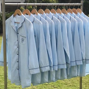 May include: A row of light blue pajama shirts hanging on wooden hangers. Each shirt has black trim on the collar, cuffs, and pockets. The shirts are button-down with a small embroidered design on the pocket.