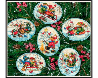 May include: Six round ornaments with a winter theme featuring snowmen, a snowman family sledding, a snowman family ice skating, a snowman roasting marshmallows by a campfire, and two snowmen decorating a Christmas tree. The ornaments are surrounded by pine branches and red berries.