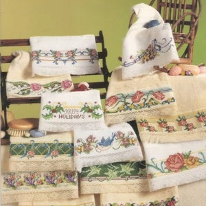May include: A collection of embroidered hand towels featuring various floral and festive designs. The towels are displayed on a wooden rack and a chair. The designs include flowers, birds, and the phrase "Happy Holidays".