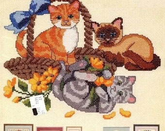 May include: Cross stitch pattern featuring three cats in a basket with flowers. The pattern includes a tabby cat, a Siamese cat, and a gray tabby cat. The pattern is framed and includes a small tag with the text "0516 7351 17".