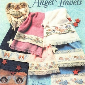 Op de afbeelding: Kruissteekpatronen voor engelhanddoeken. De patronen bevatten engelen, sterren en bloemen. De omslag van het boek bevat de titel "Angel Towels" en de auteur "by Jorja Hernandez".