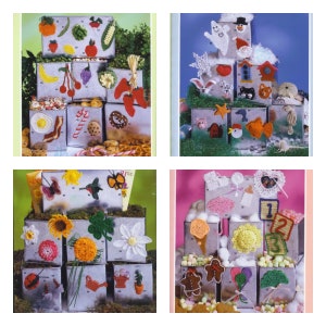 May include: Four sets of crocheted items displayed on top of metal boxes. The first set features food items like a strawberry, banana, grapes, and a red pepper. The second set features winter-themed items like a snowman, a snowflake, and a birdhouse. The third set features nature-themed items like a butterfly, a hummingbird, and a sunflower. The fourth set features baby-themed items like a pacifier, a rattle, and a baby hat.