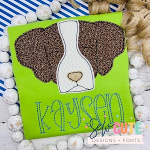 May include: A green t-shirt with an embroidered brown and white dog face and the name "Kaysen" stitched in blue thread. The text "Sew Cute Designs + Fonts" is also visible on the shirt.