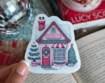 May include: A sticker featuring a pink bookstore with a red roof and a sign that reads "Bookstore". The sticker has a Christmas tree and a wreath on the door. The sticker is held by a person.