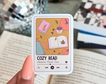 May include: A rectangular sticker with a cozy autumn theme. The sticker features illustrations of a book, pumpkins, a coffee cup, and leaves. The text "COZY READ Autumn Book Club" is displayed at the bottom, along with media player controls.