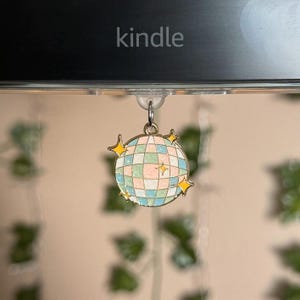 Op de afbeelding: Een gouden hanger met een roze, blauwe en witte discobal-ontwerp. De hanger heeft gele sterren eromheen en is bevestigd aan een doorzichtige plastic haak.