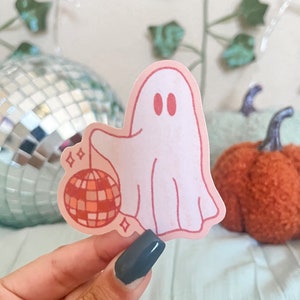 Pu&ograve; includere: Un fantasma rosa che tiene una palla da discoteca rossa e arancione con accenti di glitter.