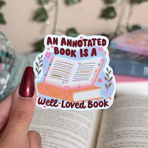 May include: A sticker with the text "An Annotated Book Is A Well-Loved Book" featuring an illustration of an open book atop a stack of books. The sticker has a white border and is decorated with floral and leaf accents.